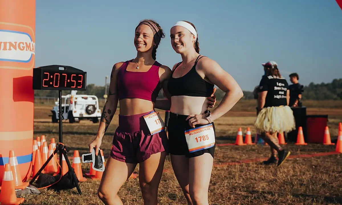 Two smiling runners posing by a race timer showing 2:07:53