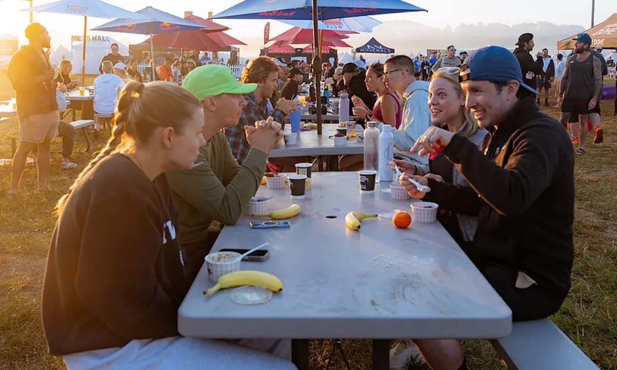 Group of people seated at long outdoor picnic table eating fruit and coffee