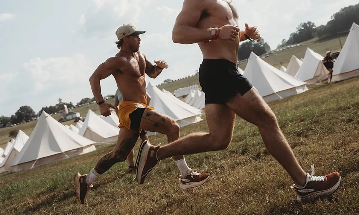 Two shirtless men sprinting across a grassy campsite with white canvas tents