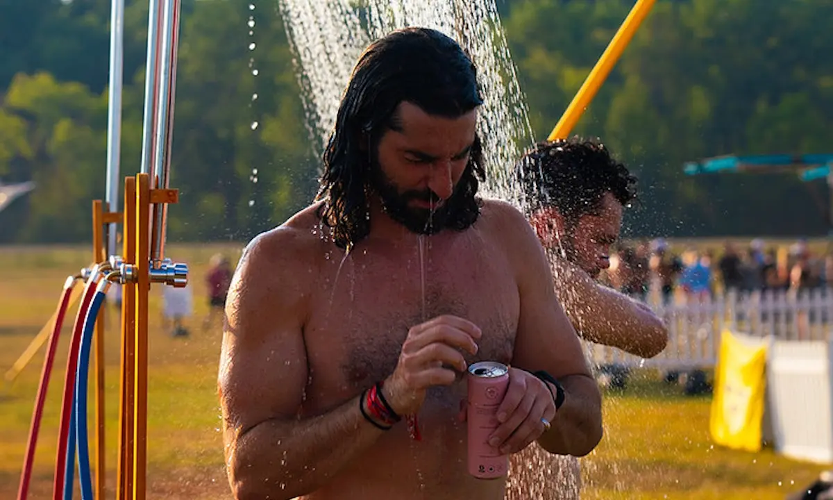 Shirtless man under outdoor shower holding a canned drink